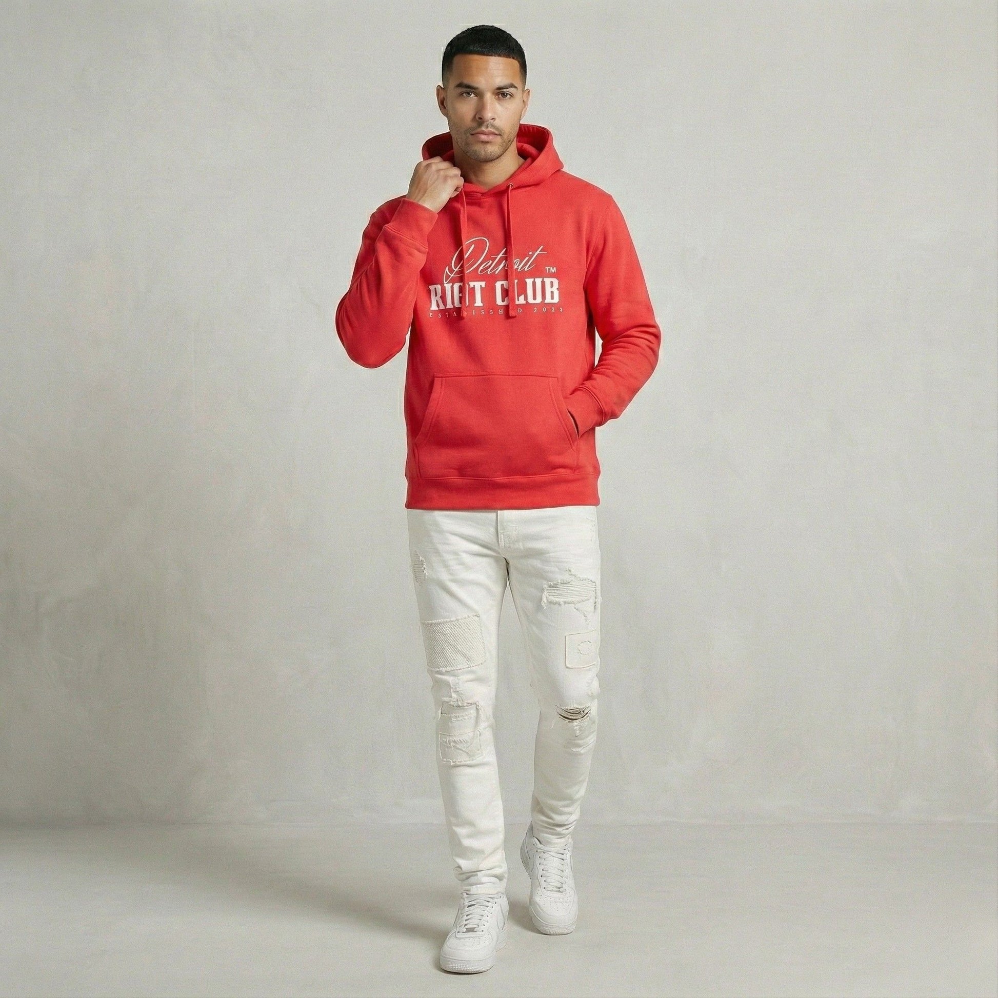 Man wearing a red hoodie with text, white pants, and white sneakers on a plain background