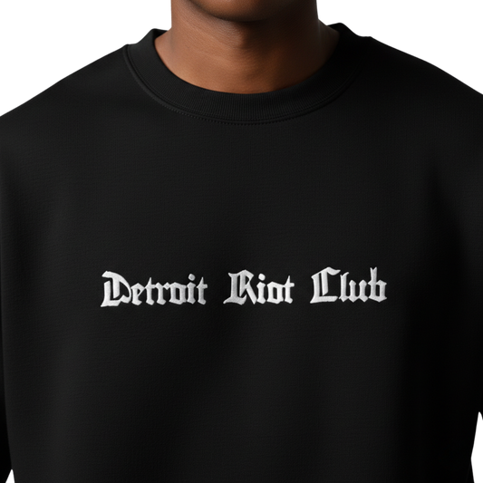 "Close-up of the male model wearing the 8.5oz Archival Crewneck hardware. Showcases premium white brand embroidery on an ultra-smooth 100% cotton face. Part of the 8.5oz archival uniform system."